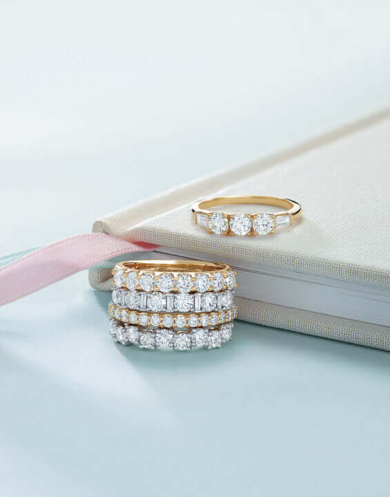 Three diamond rings on a light blue surface with a pink ribbon and beige book.