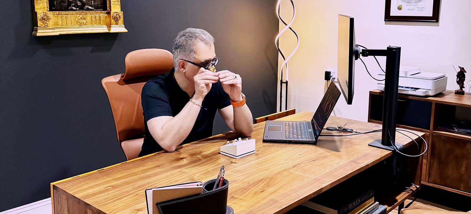 kam sitting at a desk with a laptop and computer monitor, wearing glasses and holding a small object.