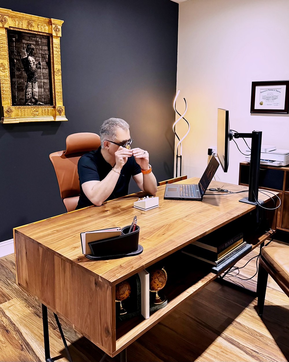 kam sitting at a wooden desk in an office setting with a laptop and computer monitor.