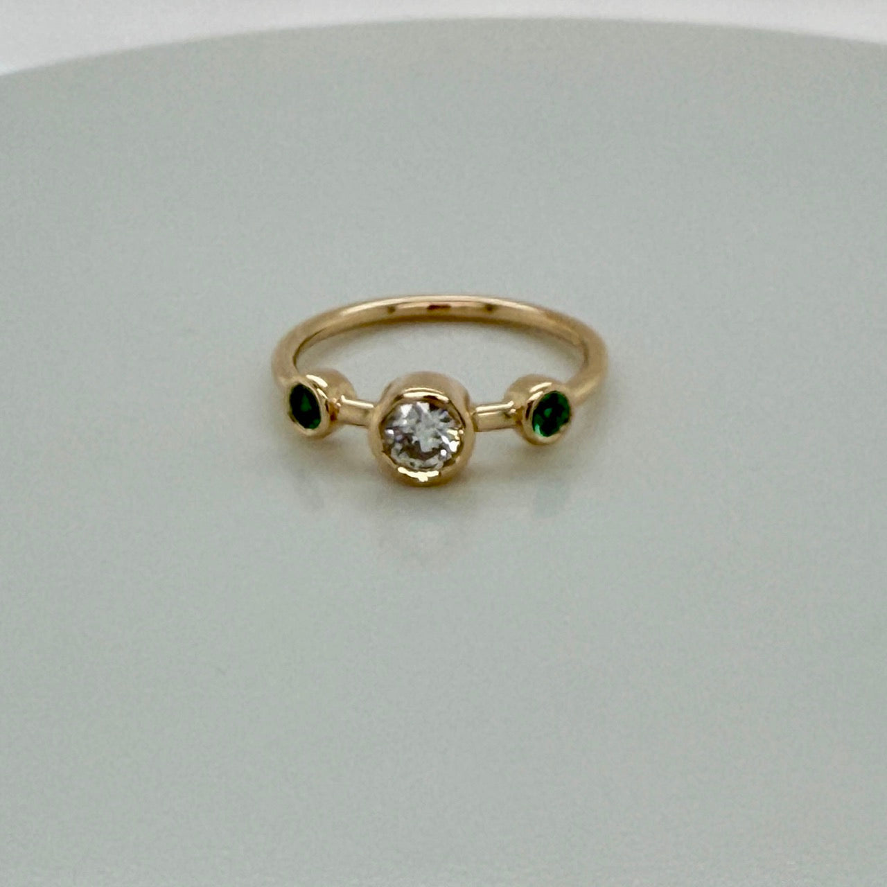 14K yellow gold three-stone ring, a round diamond bezel-set at the centre with a vivid green emerald bezel-set on either side, on a slim polished band