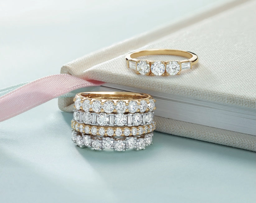 Stack of diamond rings on a light blue surface with a pink ribbon and beige book.