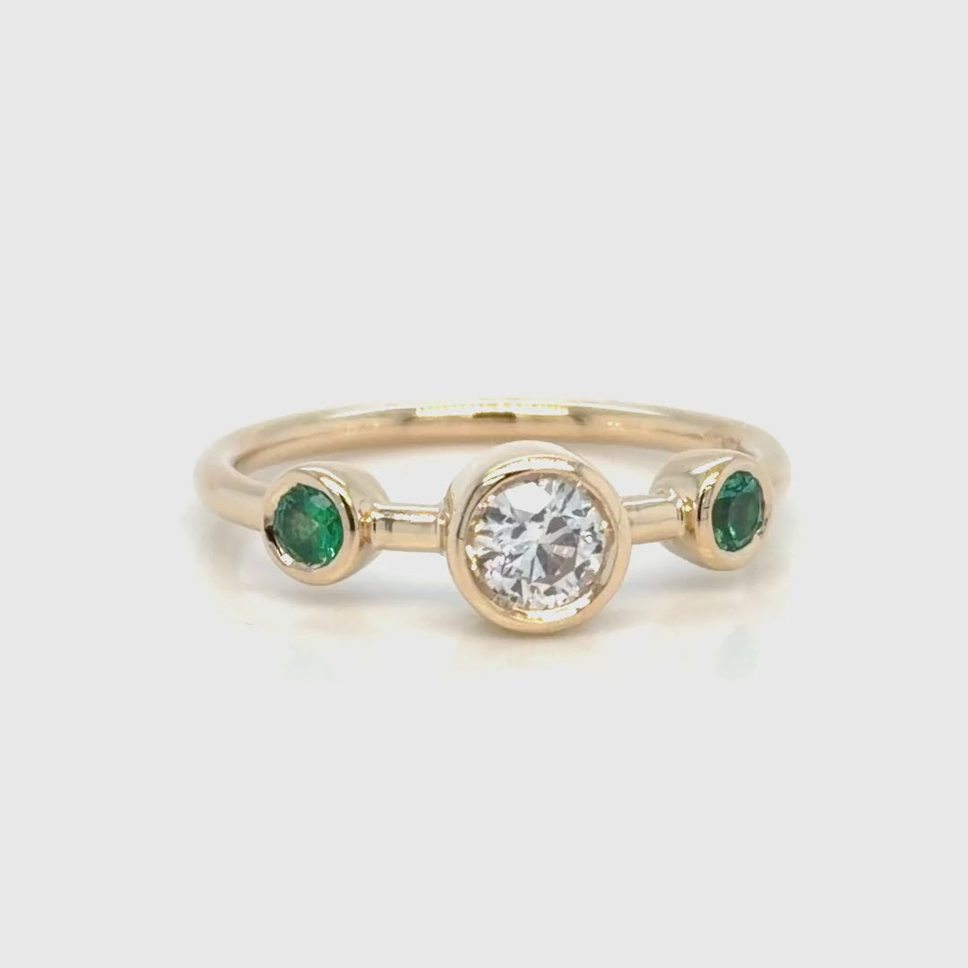 The three-stone ring turning slowly on white, showing the bezel-set round diamond at the centre and a bezel-set green emerald on each side of a slim polished 14K yellow gold band