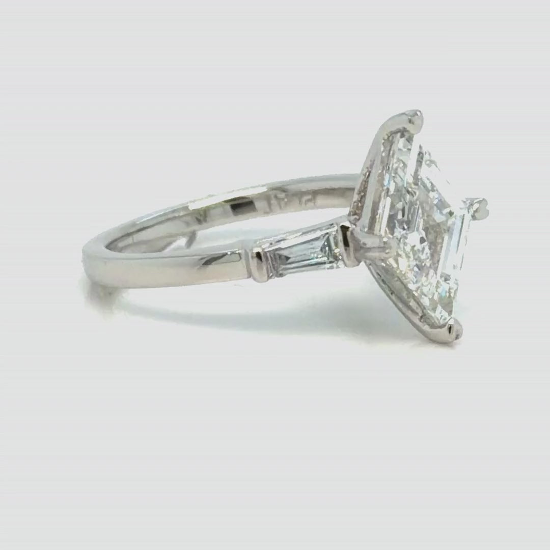 The platinum ring turning slowly from profile toward face-up, showing the lozenge-cut center held at its two points, the tapered baguettes set into the shoulders and the plain platinum band