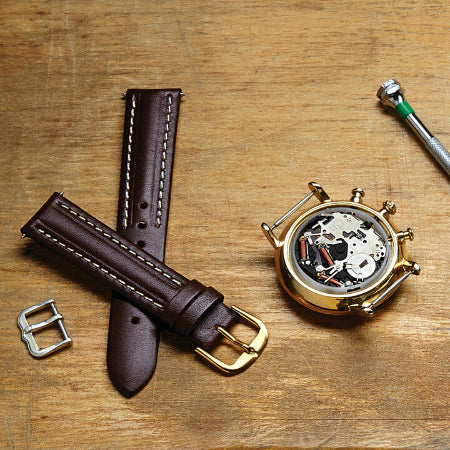 Brown leather watch strap with gold buckle on a wooden surface, alongside a gold pocket watch.